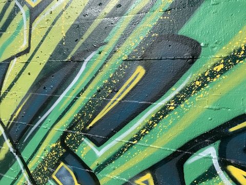 Graffiti art with bold, angular shapes and vibrant colors covers a textured wall. The design incorporates layers of green, yellow, and black with splashes of paint. The style suggests a dynamic and energetic feel, achieved through the use of sharp lines and contrasting shades.
