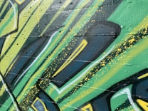Graffiti art with bold, angular shapes and vibrant colors covers a textured wall. The design incorporates layers of green, yellow, and black with splashes of paint. The style suggests a dynamic and energetic feel, achieved through the use of sharp lines and contrasting shades.