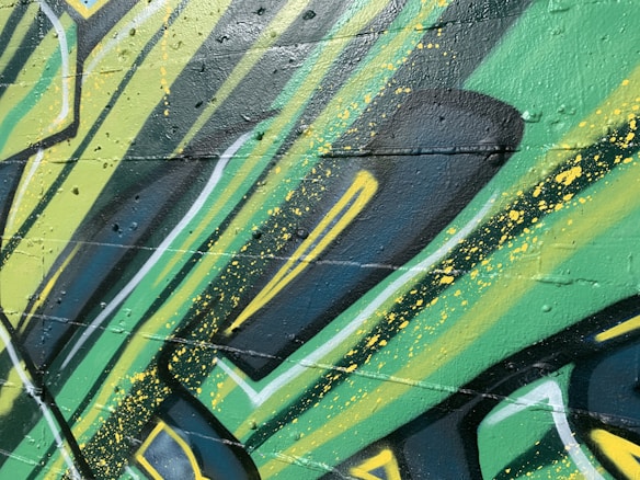 Graffiti art with bold, angular shapes and vibrant colors covers a textured wall. The design incorporates layers of green, yellow, and black with splashes of paint. The style suggests a dynamic and energetic feel, achieved through the use of sharp lines and contrasting shades.