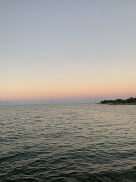 A calm sea with gentle waves under a gradient sky transitioning from soft pink to light blue, suggesting a sunset or sunrise. The horizon features a distant shoreline lined with trees and what appears to be a dock or pier extending into the water.