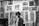 Black and white art gallery with several intricate and detailed prints displayed on the walls. The prints depict various surreal and abstract scenes, including animals, people, and imaginative designs. Two men are present in the scene, one standing and the other partially visible.