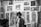 Black and white art gallery with several intricate and detailed prints displayed on the walls. The prints depict various surreal and abstract scenes, including animals, people, and imaginative designs. Two men are present in the scene, one standing and the other partially visible.