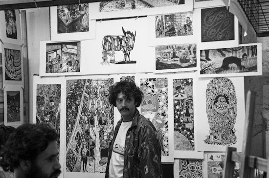 Black and white art gallery with several intricate and detailed prints displayed on the walls. The prints depict various surreal and abstract scenes, including animals, people, and imaginative designs. Two men are present in the scene, one standing and the other partially visible.