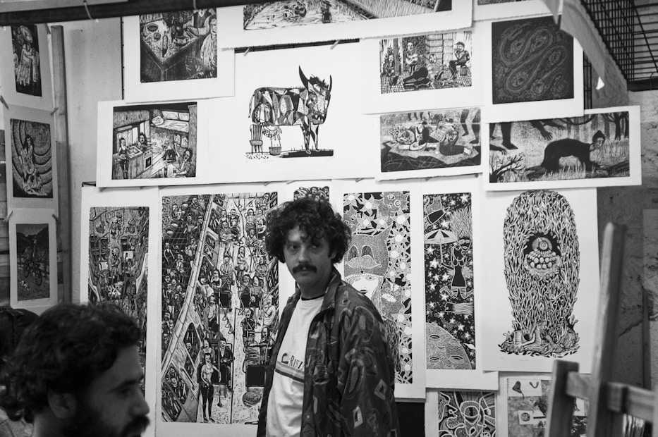 Black and white art gallery with several intricate and detailed prints displayed on the walls. The prints depict various surreal and abstract scenes, including animals, people, and imaginative designs. Two men are present in the scene, one standing and the other partially visible.