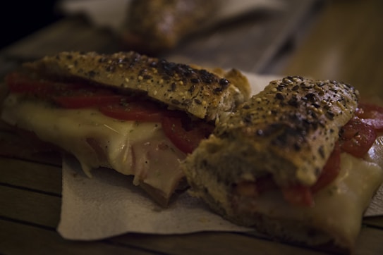 A sandwich made with seeded bread, sliced tomatoes, and melted cheese is placed on a paper napkin on a wooden surface. The sandwich appears fresh and appetizing.