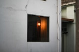 A white wall in the foreground featuring a window with a warm, glowing light bulb inside, partially illuminating the interior. There are visible wires trailing along the wall, and a partial view of another building with a light green wall and brown roof edge.