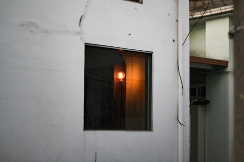 A white wall in the foreground featuring a window with a warm, glowing light bulb inside, partially illuminating the interior. There are visible wires trailing along the wall, and a partial view of another building with a light green wall and brown roof edge.