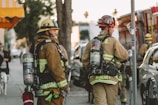 Firefighters in full gear are standing on a city street, each carrying equipment including air tanks and helmets. Their focus appears to be on a task ahead, possibly responding to an emergency. A fire truck is parked nearby, and other vehicles line the street. The scene is set in an urban environment with trees and a building canopy visible.
