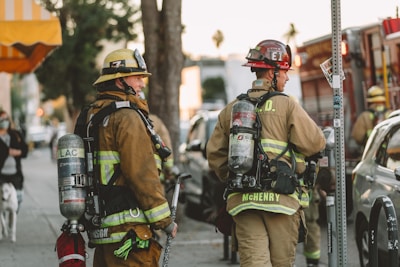 Firefighters in full gear are standing on a city street, each carrying equipment including air tanks and helmets. Their focus appears to be on a task ahead, possibly responding to an emergency. A fire truck is parked nearby, and other vehicles line the street. The scene is set in an urban environment with trees and a building canopy visible.