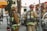 Firefighters in full gear are standing on a city street, each carrying equipment including air tanks and helmets. Their focus appears to be on a task ahead, possibly responding to an emergency. A fire truck is parked nearby, and other vehicles line the street. The scene is set in an urban environment with trees and a building canopy visible.