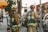 Firefighters in full gear are standing on a city street, each carrying equipment including air tanks and helmets. Their focus appears to be on a task ahead, possibly responding to an emergency. A fire truck is parked nearby, and other vehicles line the street. The scene is set in an urban environment with trees and a building canopy visible.