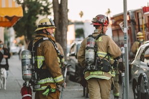 Firefighters in full gear are standing on a city street, each carrying equipment including air tanks and helmets. Their focus appears to be on a task ahead, possibly responding to an emergency. A fire truck is parked nearby, and other vehicles line the street. The scene is set in an urban environment with trees and a building canopy visible.