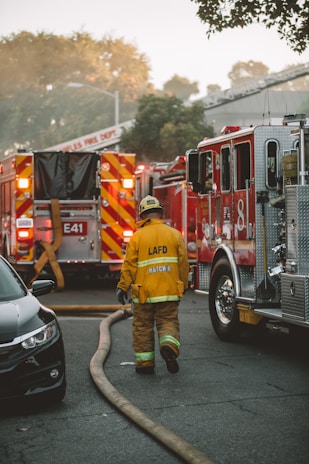 A firefighter in yellow protective gear with the initials LAFD is walking towards several fire trucks parked on a street. Thick fire hoses are spread across the road, and the background shows trees and a building under a hazy sky.
