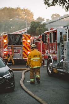 A firefighter in yellow protective gear with the initials LAFD is walking towards several fire trucks parked on a street. Thick fire hoses are spread across the road, and the background shows trees and a building under a hazy sky.