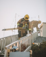 Two firefighters are clad in full gear and helmets while maneuvering on an extended firetruck ladder. They hold equipment in their hands as they perform their task. The scene appears to be set outdoors with a clear sky and some string lights in the background.