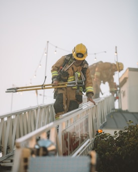 Two firefighters are clad in full gear and helmets while maneuvering on an extended firetruck ladder. They hold equipment in their hands as they perform their task. The scene appears to be set outdoors with a clear sky and some string lights in the background.