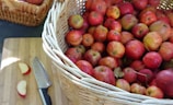 A large wicker basket filled with numerous red apples is placed on a wooden surface next to a knife and some apple slices. The apples vary slightly in color, ranging from deep red to pale yellow with red streaks. Another smaller basket filled with apples can be seen in the background.