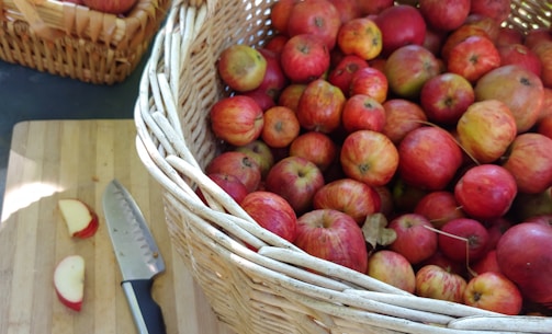 A large wicker basket filled with numerous red apples is placed on a wooden surface next to a knife and some apple slices. The apples vary slightly in color, ranging from deep red to pale yellow with red streaks. Another smaller basket filled with apples can be seen in the background.