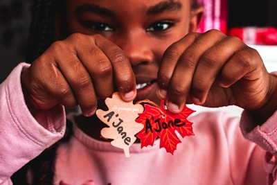 A child is holding two paper leaves in the shape of traditional autumn leaves. One leaf is brown and the other is red, both inscribed with 'Ajane'. The child's hands and part of their face are visible, with a background that includes blurred red and white elements.