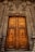 An intricately carved wooden door set within a detailed stone archway, featuring sculptures and ornate patterns. The door is tall and grand, with a deep amber color and metal accents creating a sense of historic elegance.