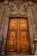 An intricately carved wooden door set within a detailed stone archway, featuring sculptures and ornate patterns. The door is tall and grand, with a deep amber color and metal accents creating a sense of historic elegance.