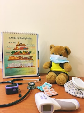 A teddy bear wearing a face mask is seated on a wooden surface beside a health guide chart depicting a food pyramid. Several medical items, including a stethoscope, blood pressure monitor, tablets, and an infrared thermometer, surround the teddy bear.