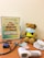 A teddy bear wearing a face mask is seated on a wooden surface beside a health guide chart depicting a food pyramid. Several medical items, including a stethoscope, blood pressure monitor, tablets, and an infrared thermometer, surround the teddy bear.