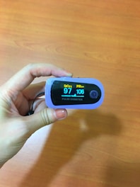A hand holds a pulse oximeter displaying SpO2 at 97% and PR at 106 bpm. The background is a wooden surface.