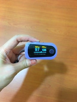 A hand holds a pulse oximeter displaying SpO2 at 97% and PR at 106 bpm. The background is a wooden surface.