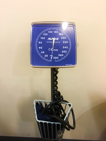 A blue analog sphygmomanometer is mounted on a stand, with a coiled tube leading to a cuff stored in a basket below. The face of the device displays a circular dial with measurements ranging from 0 to 300 mmHg.