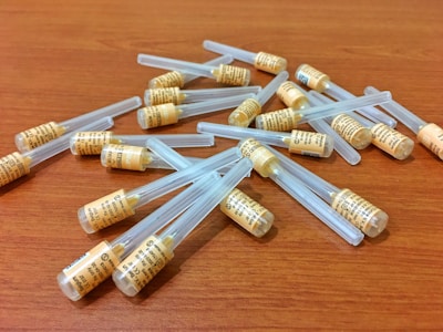 A collection of small, cylindrical containers with orange labels and transparent lids spread across a wooden surface. The containers appear to be uniform in size and shape, suggesting they are medical or laboratory-related.