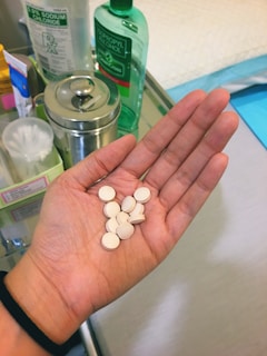 A hand holding several round white tablets, resting above a surface with various medical supplies such as a bottle of isopropyl alcohol, a container of sodium chloride, a metal canister, and other personal care items.