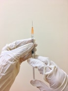 A pair of hands wearing white latex gloves is holding a syringe with an orange cap. The syringe is filled with a small amount of liquid and the background is a plain, light-colored wall.