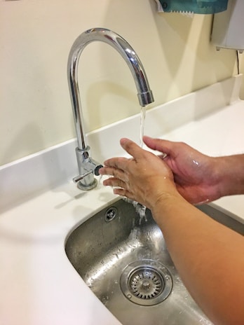 Hands are being washed under a running faucet in a bathroom sink. Water is flowing from the metallic, curved faucet onto the hands. The sink and counter are clean and white, showcasing a tidy environment.