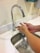 Hands are being washed under a running faucet in a bathroom sink. Water is flowing from the metallic, curved faucet onto the hands. The sink and counter are clean and white, showcasing a tidy environment.