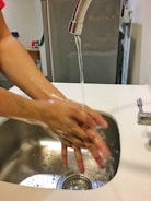 A person is seen washing their hands under a running faucet in a kitchen sink. Soap bubbles are visible, indicating the hands are being thoroughly cleaned. The background contains kitchen appliances and items, providing a domestic setting.