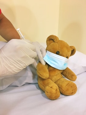 A teddy bear wearing a blue surgical mask is seated on a white pillow. A person's hands, wearing gloves, are administering a vaccine with a syringe to the teddy bear.