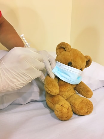 A teddy bear wearing a blue surgical mask is seated on a white pillow. A person's hands, wearing gloves, are administering a vaccine with a syringe to the teddy bear.