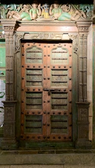 An ornate, wooden door with intricate carvings and embellishments. The door features a series of decorative studs and panels, flanked by elaborately carved columns. Above the doorway, there are detailed sculptural figures in vibrant colors, portraying seated individuals in traditional attire.