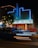 A vintage theater building illuminated with blue neon lights at night. The marquee displays the message 'Movies Are Forever, We'll See You Soon.' The architecture features a retro style with brick walls and a vertical sign. Light trails from passing cars are visible in the foreground, adding a dynamic element to the scene. A tree with string lights is on the left side.