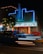 A vintage theater building illuminated with blue neon lights at night. The marquee displays the message 'Movies Are Forever, We'll See You Soon.' The architecture features a retro style with brick walls and a vertical sign. Light trails from passing cars are visible in the foreground, adding a dynamic element to the scene. A tree with string lights is on the left side.