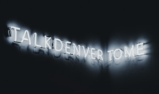Neon letters forming the phrase 'TALK DENVER TO ME' glowing against a dark background, creating a striking contrast.