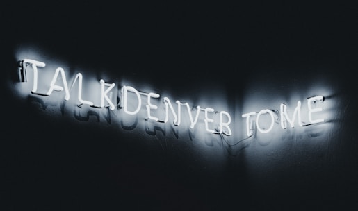 Neon letters forming the phrase 'TALK DENVER TO ME' glowing against a dark background, creating a striking contrast.