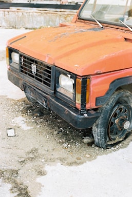 A rusted, abandoned car with damaged bodywork and missing wheel parts. The car is parked on a rough, gravelly surface with visible rust and paint deterioration. Surrounding debris and a worn-down environment contribute to a sense of neglect.