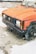 A rusted, abandoned car with damaged bodywork and missing wheel parts. The car is parked on a rough, gravelly surface with visible rust and paint deterioration. Surrounding debris and a worn-down environment contribute to a sense of neglect.
