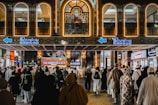 A busy marketplace scene with numerous people walking in front of a building adorned with bright lights and intricate patterns. The shop signs are illuminated, with the prominent 'Bin Dawood' sign in both English and Arabic. The architecture features arched windows and decorative tile work.
