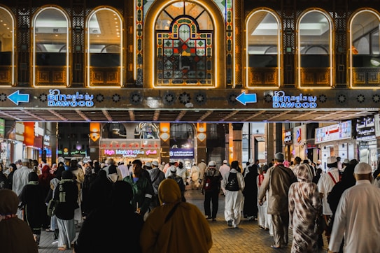 A busy marketplace scene with numerous people walking in front of a building adorned with bright lights and intricate patterns. The shop signs are illuminated, with the prominent 'Bin Dawood' sign in both English and Arabic. The architecture features arched windows and decorative tile work.