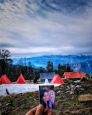 A picturesque campsite set against a backdrop of majestic mountains and a partly cloudy sky. Several bright red and orange tents are pitched on a snowy ground, with trees scattered around. In the foreground, a hand holds a photograph of two smiling women.