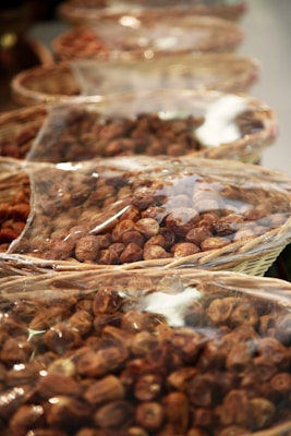 Several wicker baskets filled with nuts are lined up in a row. Each basket is covered with a clear plastic sheet, giving a glimpse of the contents packed inside. The nuts appear to be of the same type, uniformly brown and small in size.