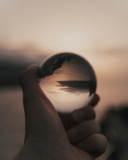 A hand holding a crystal ball that reflects an upside-down image of a landscape at sunset, with a cloudy sky and a distant landmass across a body of water. The background appears blurred, accentuating the clarity within the crystal ball.